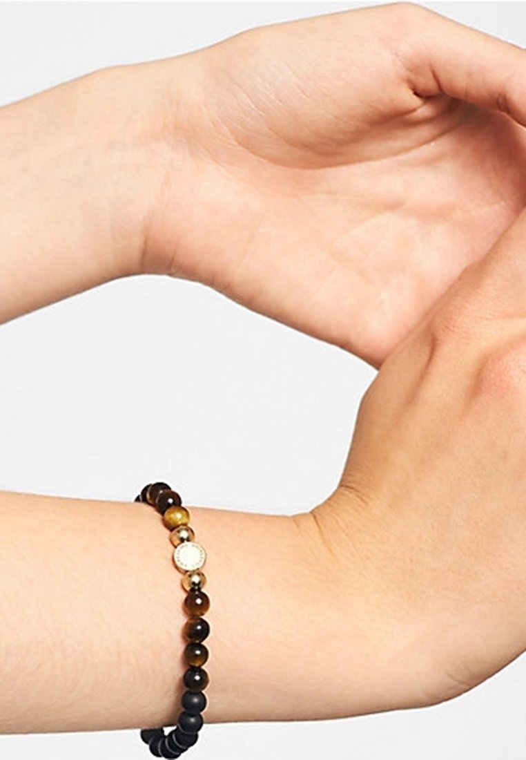 Bras avec un bracelet de perles comprenant des perles noires, marron et dorées avec un petit charme rond en or, sur un fond clair uni.