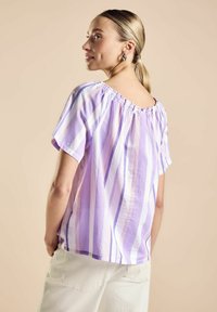 Blusa a righe lilla con maniche corte e scollo arricciato. Tessuto morbido con vestibilità comoda. Indossata con pantaloni bianchi.