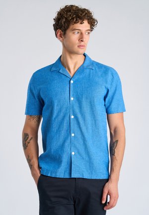 Mężczyzna o kręconych włosach, ubrany w niebieską, krótkorękawową koszulę zapinaną na guziki oraz czarne spodnie, patrzący w bok z rękami w kieszeniach.