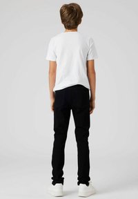 T-shirt blanc à manches courtes, jean noir slim, et baskets blanches ; vu de dos contre un fond neutre.