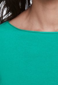 Gros plan sur le cou et l'épaule d'une personne portant un t-shirt col rond vert avec des cheveux foncés partiellement visibles.