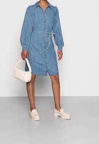 Denim skjortklänning med långa puffärmar, knäppning fram, bälte i midjan och knäslång nederkant. Buren med beiga platå-tofflor och en flätad handväska.