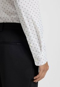 Camicia bianca con un piccolo motivo ripetuto di strisce rosse e nere, con polsini chiusi da bottoni e tessuto liscio, abbinata a pantaloni neri strutturati.