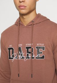 Felpa cappuccio marrone realizzata in un tessuto morbido con cappuccio regolabile. Nella parte anteriore presenta una grande scritta "DARE" in bianco e nero, con un testo più piccolo sopra.