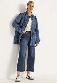 Farkkutakki etutaskuilla, yhdistettynä tumman farkun leveisiin housuihin ja valkoisiin sneaker-kengiin mustilla raitoilla. Yksinkertainen, rento ilme.