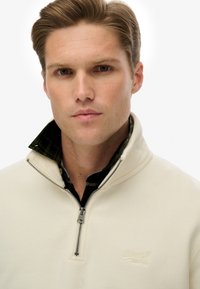 Lichtcrème ritsfleece met een staande kraag, voorzien van een gestructureerd patroon in de binnenlaag en een klein geborduurd logo op de borst.