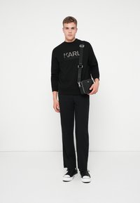 Mladý muž v čiernej mikine Karl Lagerfeld, čiernych nohaviciach, čierno-bielych teniskách, drží čiernu crossbody tašku na bielom pozadí.