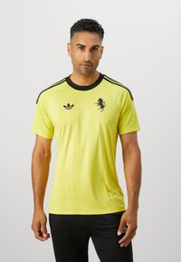 Žltý športový dres s čiernymi pruhmi na ramenách, s čiernym logom koňa a emblémom Adidas trefoil na ľavej hrudi.