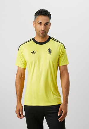 Žltý športový dres s čiernymi pruhmi na ramenách, s čiernym logom koňa a emblémom Adidas trefoil na ľavej hrudi.