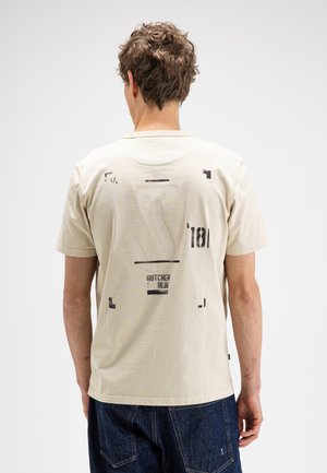 Man met kort krullend haar draagt een offwhite T-shirt met een abstracte zwarte print en tekst "Butcher Blue 181" op de achterkant, gecombineerd met donkerblauwe spijkerbroek.