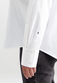 Primo piano di una mano di una persona che indossa una camicia bianca a maniche lunghe con un ricamo di un piccolo fiore nero sul polsino, abbinata a pantaloni scuri.
