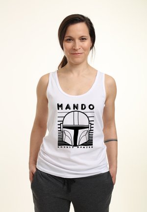 Top sin mangas blanco con texto gráfico negro "MANDO" y un diseño de casco ilustrado, combinado con pantalones gris oscuro. Textura de tela suave.