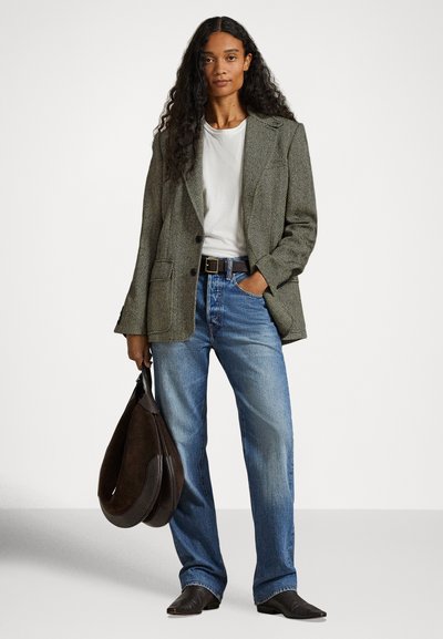 Kobieta z długimi kręconymi włosami, ubrana w wzorzysty żakiet, białą koszulkę, niebieskie jeansy, czarny pasek, czarne buty i trzymająca dużą ciemnobrązową torbę.