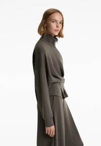 Olive green sweatshirt with high collar and zip, loose fit, long sleeves, and tied detail at the waist, made of soft fabric.