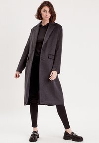 Langer, grauer Wollmischmantel mit tailliertem Design, zwei Knöpfen und seitlichen Taschen. Getragen über einem schwarzen Oberteil und Hose.