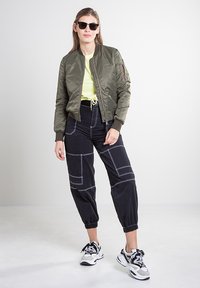 Olivengroen bomberjack met geribbelde manchetten, felgeel shirt, zwarte taps toelopende broek met witte stiksels en sneakers met patroon.