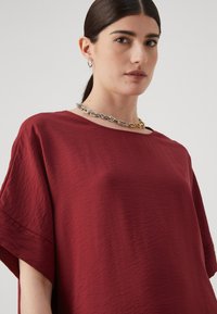 Burgundy oversized topp med en strukturerad tyg, rund halsringning och vida ärmar, matchad med ett lager av silver- och guldkedjor.