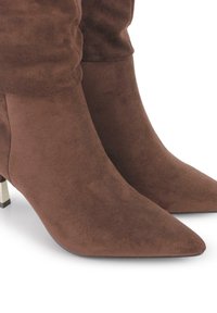 Botas de ante marrón hasta el tobillo con punta afilada y un tacón bajo acentuado con metal, que presentan un eje recogido para añadir textura.