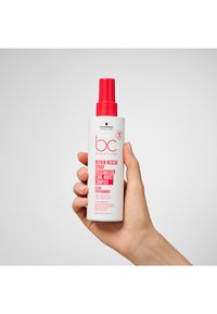 Flacon pulvérisateur blanc avec un embout rouge, étiqueté "BC Bonacure Repair Rescue Spray Conditioner", tenu dans une main sur un fond clair.