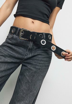 Cintura in suede nera con multiple occhielli in argento e una fibbia metallica lucida, indossata sopra jeans grigio scuro su uno sfondo neutro.