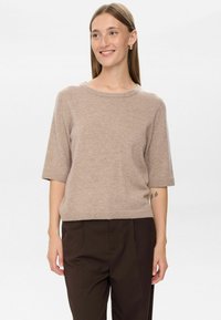 Leende kvinna med rakt hår bär en beige kortärmad tröja, mörkbruna byxor och guldcreoler framför en helt vit bakgrund.