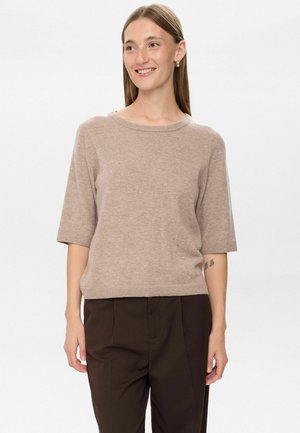 Femme souriante avec les cheveux raides portant un pull beige à manches courtes, un pantalon marron foncé et des boucles d'oreilles en or sur fond blanc uni.