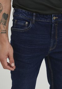 Tamno plave traperice slim fit kroja, s detaljnim šavovima, prednjim džepovima i vidljivim kopčanjem na dugme. Glatka tekstura i minimalno izbljeđivanje.