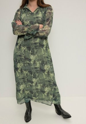 Donna con un lungo vestito verde a motivi con maniche trasparenti, braccia incrociate, indossando stivali alla caviglia neri con borchie, in piedi davanti a uno sfondo semplice.