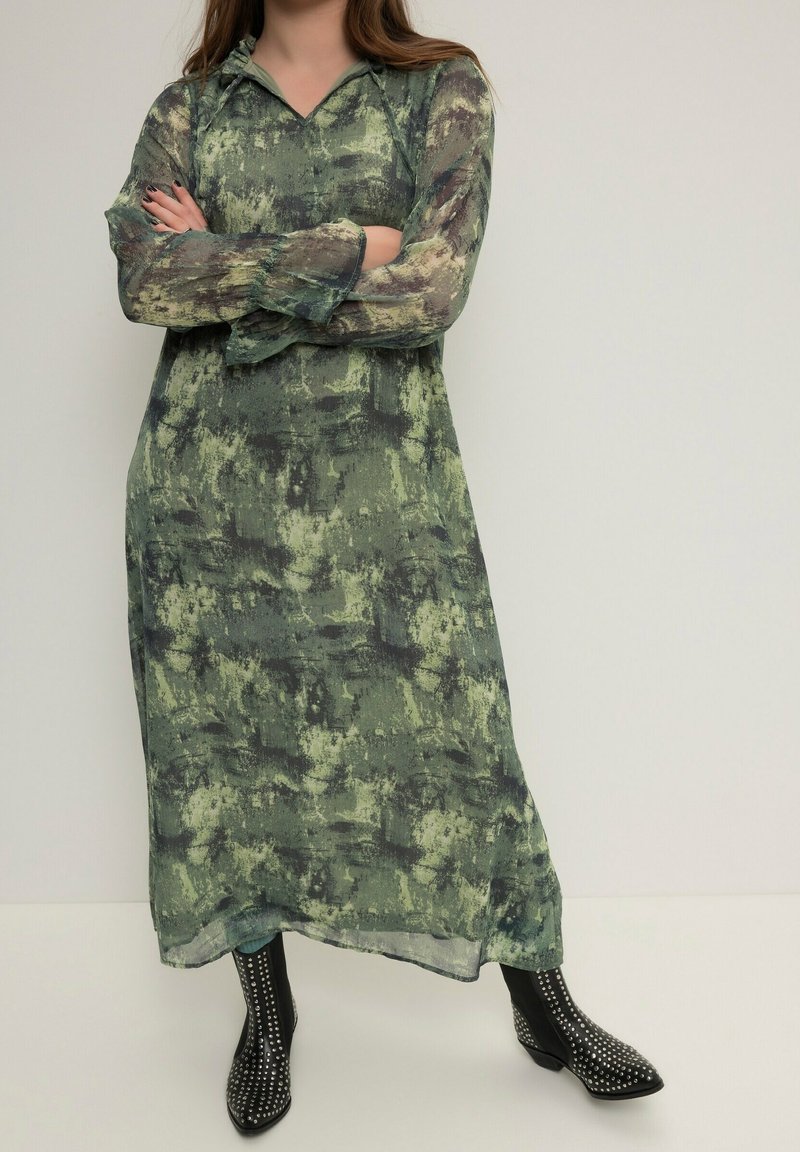 Mujer con un vestido largo verde con estampado y mangas transparentes, brazos cruzados, llevando botines negros con tachuelas, de pie frente a un fondo liso.