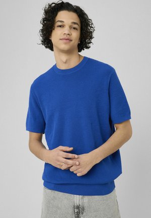 Jeune homme aux cheveux bouclés portant une chemise en maille à manches courtes bleue et un pantalon gris clair, debout devant un fond gris uni.