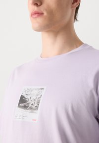 Light lavender short-sleeve t-shirt featuring a rectangular black-and-white graphic of a dog swimming, with "HUGO" in red text at the bottom.