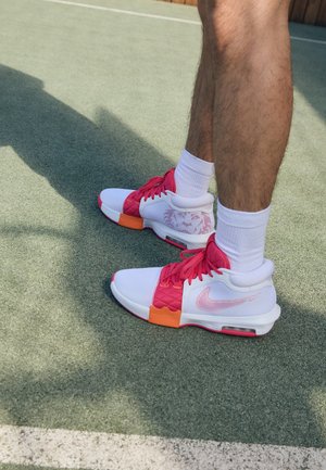 Weiße und rosa Sportschuhe mit orangefarbenen Akzenten, Netzpaneelen, strukturierten Oberflächen und sichtbarem Nike-Logo, vor einem grünen Platz.