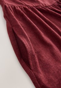 Bordeauxkleurig kledingstuk met zachte, gestructureerde stof, een ontspannen ontwerp en een geplooide taille. Zichtbare stiksels langs de naden.