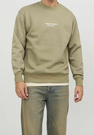 Man wearing olive green sweatshirt with white "Originals Studio" text and faded blue jeans, standing against a plain light background.