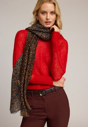 Vaaleatukkainen nainen pukeutunut punaiseen neulepaitaan, ruskeisiin housuihin, joissa on musta vyö, ja leopardikuvioiseen huiviin, poseeraa käsi leukaa vasten neutraalilla taustalla.