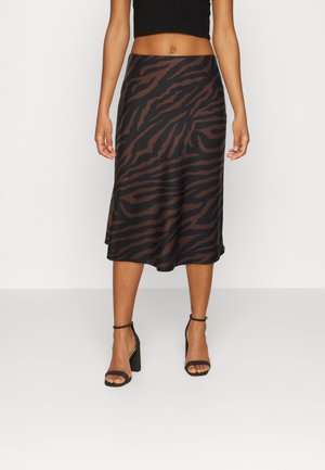 Kobieta ubrana w brązowo-czarną spódnicę midi w wzór zebry, czarny krótki top oraz czarne sandały na blokowym obcasie z paskiem wokół kostki, stojąca na białym tle.