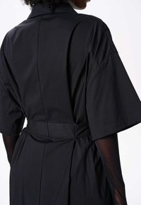 Czarna sukienka z krótkimi, szerokimi rękawami, kołnierzykowym dekoltem i związanym w talii pasem. Posiada gładką teksturę oraz delikatne marszczenia z tyłu.