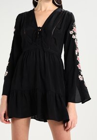 Svart långärmad klänning med V-ringning, blommig brodyr på ärmarna, en åtsittande korsett och en utsvängd, tärnad fåll. Mjuk tygkänsla.