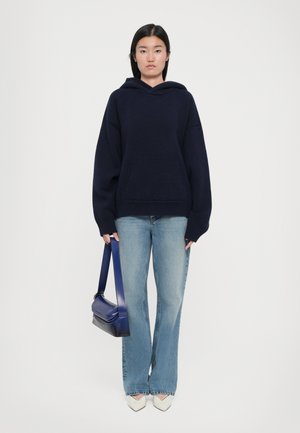 Navy hooded sweater with a pouch pocket, worn with light blue flared jeans. Model carries a small dark blue handbag and wears white heels.