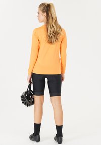 Långärmad orange träningsovershirt i kombination med svarta cykelshorts. Modellen håller en svart hjälm och har på sig svarta cykelskor och strumpor.