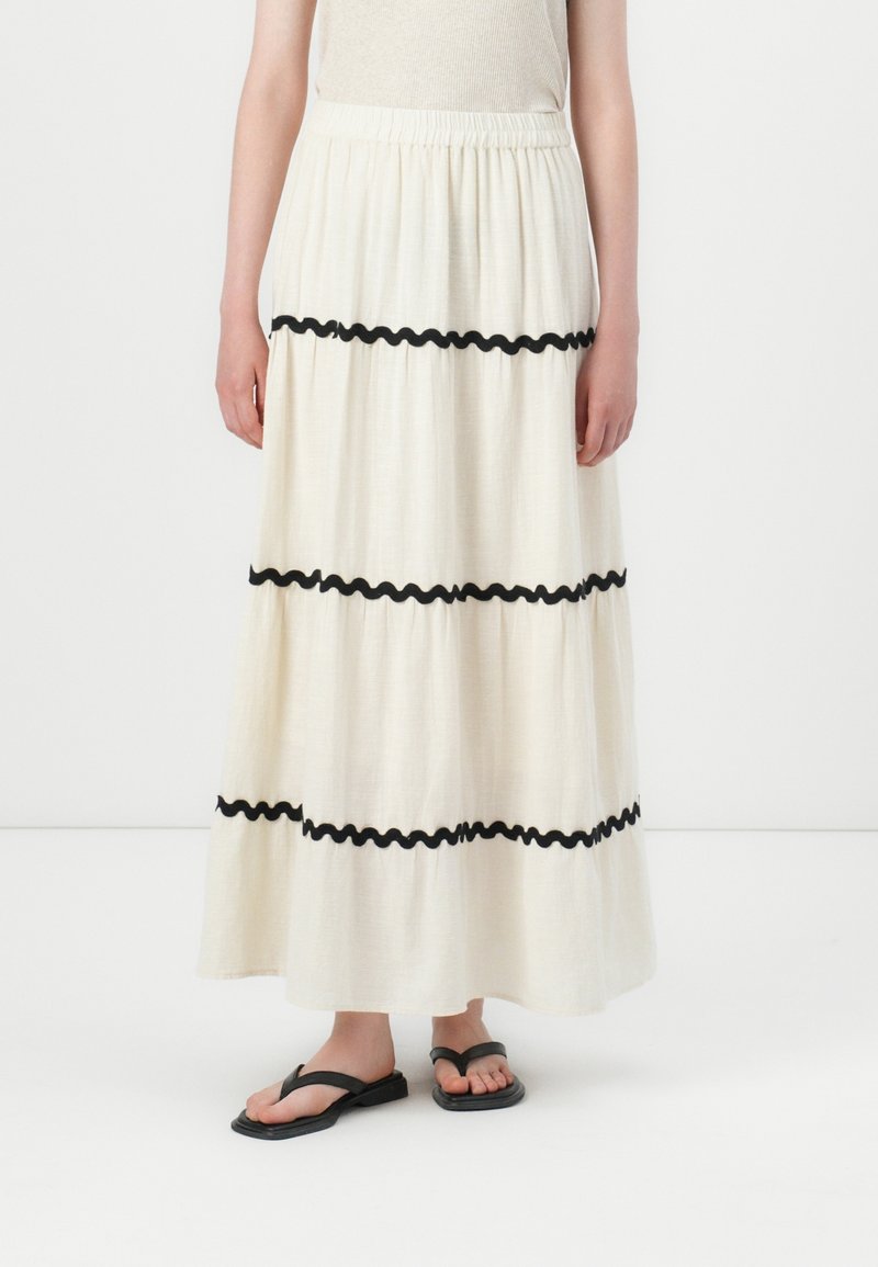 Femme portant une longue jupe crème avec trois bandes horizontales noires ondulées, associée à un haut beige et des tongs noires.
