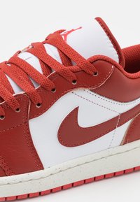 Rode en witte sneaker van leer en mesh materialen. Uitgerust met opvallende rode veters, een gestructureerde buitenzool en een kenmerkend logo aan de zijkant.