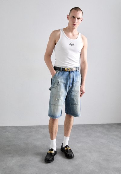 Kopott kék árnyalatú farmer rövidnadrág, nagy zsebekkel és kopott részletekkel. Egy fehér ujjatlan pólóval és fekete slip-on cipővel párosítva.