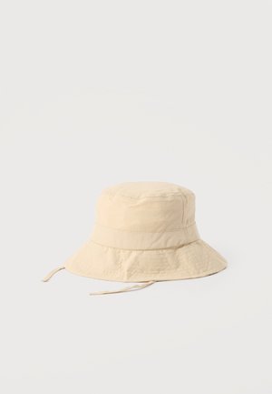 Sombrero tipo bucket beige con ala ancha y correas para la barbilla, hecho de tela ligera, exhibido sobre un fondo blanco liso.