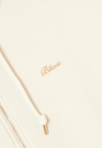 Helles beige Zip-Up Sweatshirt mit goldfarbener, gestickter "Blkvrs"-Logoplatzierung, weißen Kordeln und metallicfarbenem Reißverschluss. Glatte Stofftextur.