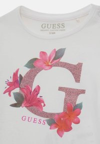 Hvid økologisk bomulds T-shirt med et stort glitrende pink bogstav "G" omgivet af blomsterdesigns i pink og grøn. Størrelse 3/6M.