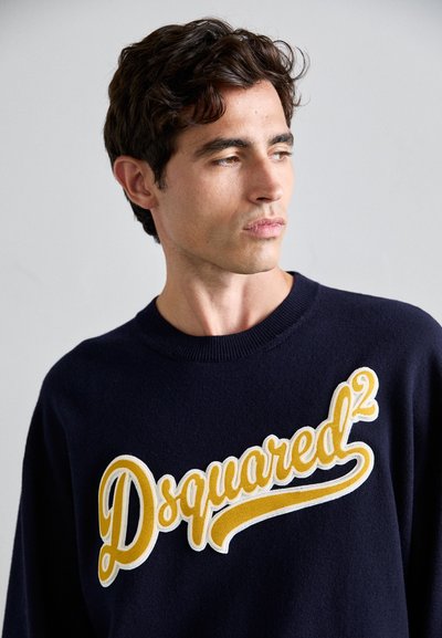 Tengerészkék kötött pulóver, amelyen egy nagy, sárga és fehér hímezett logó látható, rajta a "Dsquared²" felirattal. Klasszikus környakú fazon.