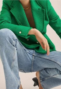 Zielony, strukturalny żakiet z podwójnymi guzikami, połączony z jasnoniebieskimi, oversize'owymi dżinsami. Widoczne czarne szpilki.