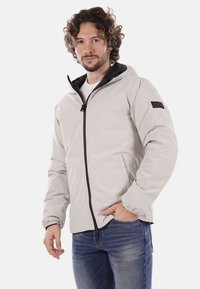 Chaqueta exterior gris claro con cremallera negra y capucha, con un acabado texturizado y un detalle en negro en la manga. Llevada sobre una camisa blanca.