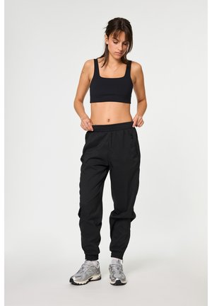Femme ajustant un pantalon de jogging noir, portant un crop top noir et des baskets argentées sur un fond clair uni.